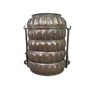 May include: Antique copper-colored food carrier with four stacked, round compartments. The carrier has a handle and is made of metal. The compartments have a decorative, textured design. The carrier is likely used for transporting food.