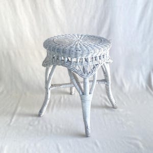 May include: A white wicker stool with a round seat and three curved legs. The stool has a woven design on the seat and a decorative pattern around the edge. The stool is painted white and is set against a white background.