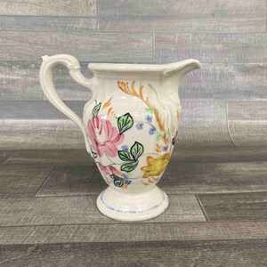 May include: A cream-colored ceramic pitcher featuring a hand-painted floral design with large pink roses, green leaves, and yellow and orange accents. The vessel has a curved handle, a flared rim with a pouring spout, and a circular pedestal base.