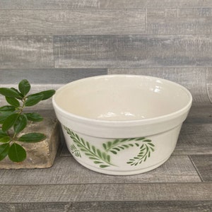 May include: A white ceramic bowl with a green leafy design. The bowl is empty and sitting on a wood-grain surface.