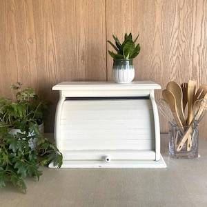 May include: A white wooden bread box with a roll-top door and a small round knob. The bread box is positioned between a green leafy plant on the left and a clear glass container filled with wooden spoons and metal whisks on the right.