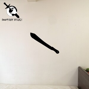May include: Black silhouette of a machete on a white wall. The machete is angled diagonally from the top left to the bottom right. The wall is plain white with no other objects or markings. The image is a product listing for a wall decal.
