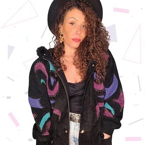 Può includere: Giacca in camoscio nera con disegni astratti viola, turchese e magenta. La giacca ha borchie dorate e una chiusura con zip. La persona indossa un cappello nero e orecchini dorati. Stile vintage.