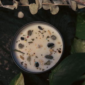 Può includere: Un barattolo di candela rotondo nero riempito di cera bianca cremosa. La cera è decorata con piccole pietre, glitter ed erbe secche. La candela è circondata da foglie verdi ed eucalipto secco.