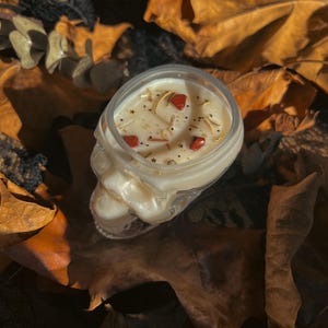 Puede incluir: Un portavelas con forma de calavera lleno de una vela blanca cremosa. La vela está decorada con pequeñas piedras rojas, estrellas doradas y otros elementos decorativos. La vela se encuentra sobre un lecho de hojas secas.