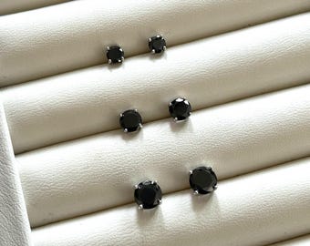 May include: A set of six black stud earrings displayed on a white velvet earring display. The earrings are arranged in three pairs, with each pair featuring a different size of black round stone.