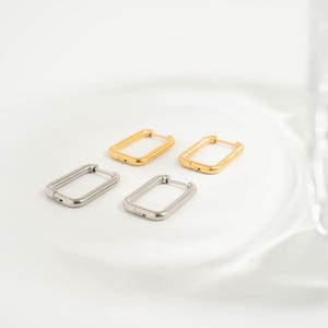 May include: Four rectangular hoop earrings, two in gold and two in silver, are displayed on a white surface. The earrings have a simple, modern design with a hinged closure. The gold earrings have a shiny finish, while the silver earrings have a matte finish.