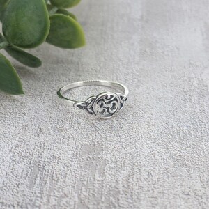 May include: A silver ring with an Om symbol in the center. The ring has a delicate design with a circular emblem and intricate details. The ring is set against a textured, light gray surface, with green leaves in the background.