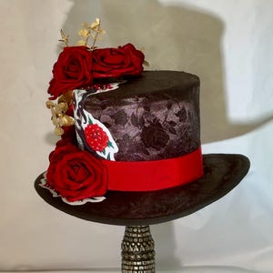 May include: A dark brown top hat featuring a textured floral pattern, adorned with a vibrant red ribbon band and a cluster of deep red artificial roses. A decorative graphic element with a skull motif is visible on the side of the hat crown.