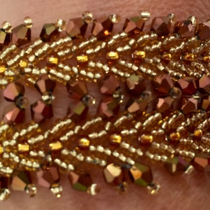May include: A gold and copper beaded bracelet with a woven design. The bracelet is made with small, faceted beads in a variety of shades of gold and copper.