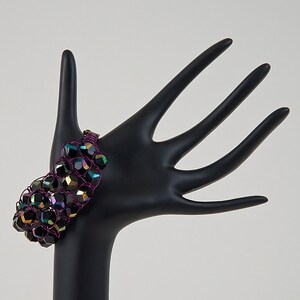 Könnte beinhalten: Ein schwarzes und schillerndes Perlenarmband mit einem violetten Drahtgeflecht. Das Armband wird auf einem schwarzen Handmodell präsentiert.