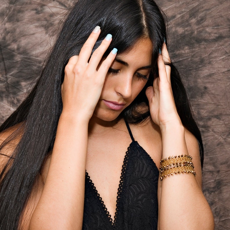May include: A woman with long black hair is wearing a black lace top and a gold bracelet. She is looking down and has her hands on her face.