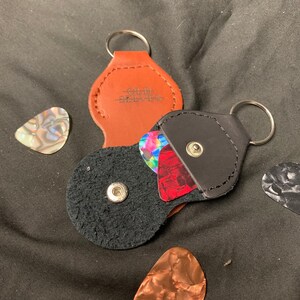 May include: Two leather guitar pick holders with keychains. One is brown with the text "Old Slivess" and the other is black. Both holders have a snap closure and are filled with guitar picks.