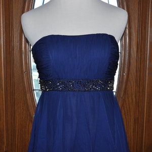 Puede incluir: Un vestido de fiesta azul marino sin tirantes. El vestido presenta un cintur&oacute;n con cuentas y una falda fluida. El corpi&ntilde;o est&aacute; fruncido y el vestido se exhibe en un maniqu&iacute;. El fondo es una puerta de madera con una ventana.