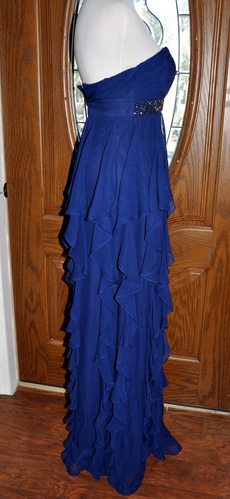 Puede incluir: Un vestido largo azul real sin tirantes con una falda con volantes en capas. El corpi&ntilde;o tiene un adorno de cuentas en la cintura. El vestido se exhibe en un maniqu&iacute;.