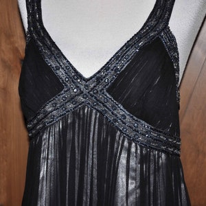 Puede incluir: Un vestido de noche negro con escote en V y tirantes finos. El corpi&ntilde;o y los tirantes est&aacute;n adornados con abalorios plateados. La falda tiene un dise&ntilde;o fluido y plisado. El vestido se muestra en un maniqu&iacute; sobre un fondo de paneles de madera.