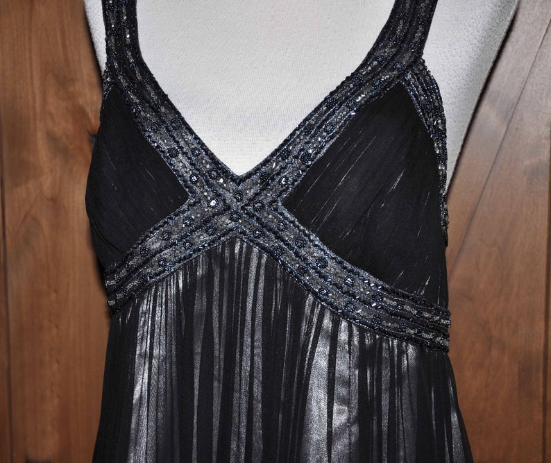 Puede incluir: Un vestido de noche negro con escote en V y tirantes finos. El corpi&ntilde;o y los tirantes est&aacute;n adornados con abalorios plateados. La falda tiene un dise&ntilde;o fluido y plisado. El vestido se muestra en un maniqu&iacute; sobre un fondo de paneles de madera.