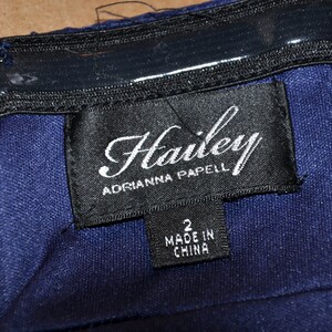 Puede incluir: Primer plano de un vestido azul marino con una etiqueta negra. La etiqueta dice "Hailey ADRIANNA PAPELL" e incluye la talla "2" y "MADE IN CHINA". El vestido tiene una cremallera.
