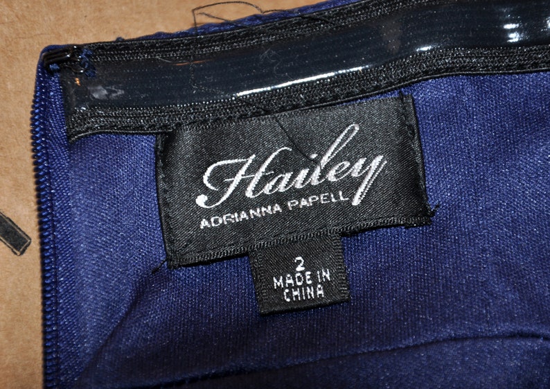 Puede incluir: Primer plano de un vestido azul marino con una etiqueta negra. La etiqueta dice "Hailey ADRIANNA PAPELL" e incluye la talla "2" y "MADE IN CHINA". El vestido tiene una cremallera.