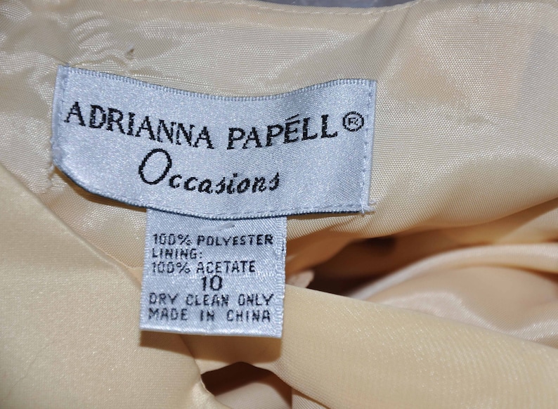 K&ouml;nnte beinhalten: Ein wei&szlig;es Etikett mit schwarzem Text, auf dem "ADRIANNA PAPELL Occasions" und "100% POLYESTER FUTTER: 100% ACETAT 10 NUR CHEMISCHE REINIGUNG HERGESTELLT IN CHINA" steht.