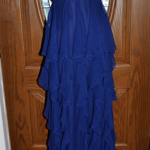 Puede incluir: Vestido de noche largo y sin tirantes en un vibrante tono azul. El corpi&ntilde;o est&aacute; fruncido y la cintura est&aacute; acentuada con una banda de cuentas. La falda presenta m&uacute;ltiples capas de volantes fluidos, creando una silueta dram&aacute;tica.