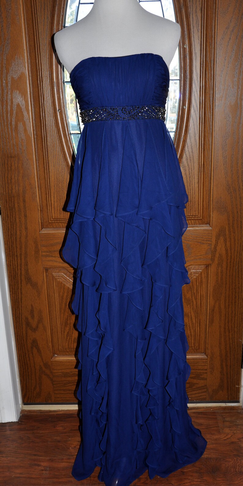 Puede incluir: Vestido de noche largo y sin tirantes en un vibrante tono azul. El corpi&ntilde;o est&aacute; fruncido y la cintura est&aacute; acentuada con una banda de cuentas. La falda presenta m&uacute;ltiples capas de volantes fluidos, creando una silueta dram&aacute;tica.