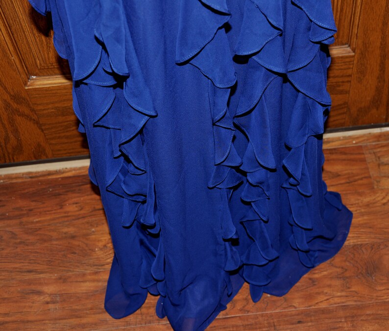 Puede incluir: Primer plano de un vestido azul real fluido con volantes en capas. Los volantes caen en cascada sobre la prenda, creando una apariencia texturizada y elegante. El vestido parece estar hecho de una tela ligera, adecuada para ocasiones formales.