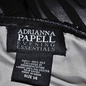 Puede incluir: Primer plano de una etiqueta negra de un vestido Adrianna Papell Evening Essentials. La etiqueta es negra con texto blanco, incluyendo la marca, la composici&oacute;n de la tela y la talla 14. El vestido parece ser negro con forro plateado.