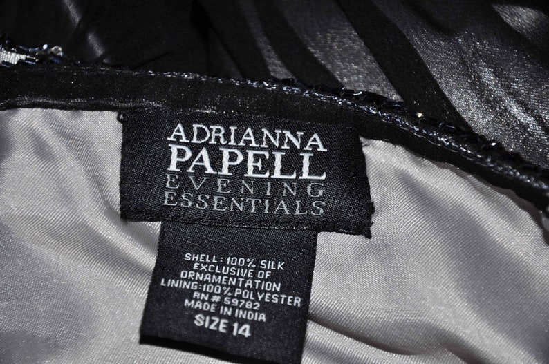 Puede incluir: Primer plano de una etiqueta negra de un vestido Adrianna Papell Evening Essentials. La etiqueta es negra con texto blanco, incluyendo la marca, la composici&oacute;n de la tela y la talla 14. El vestido parece ser negro con forro plateado.