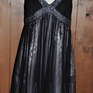 Puede incluir: Vestido de noche negro sobre un maniqu&iacute;. El vestido presenta un escote en V y tirantes adornados con cuentas plateadas. La falda es plisada y tiene un dobladillo con cuentas. El vestido es de tejido transparente.