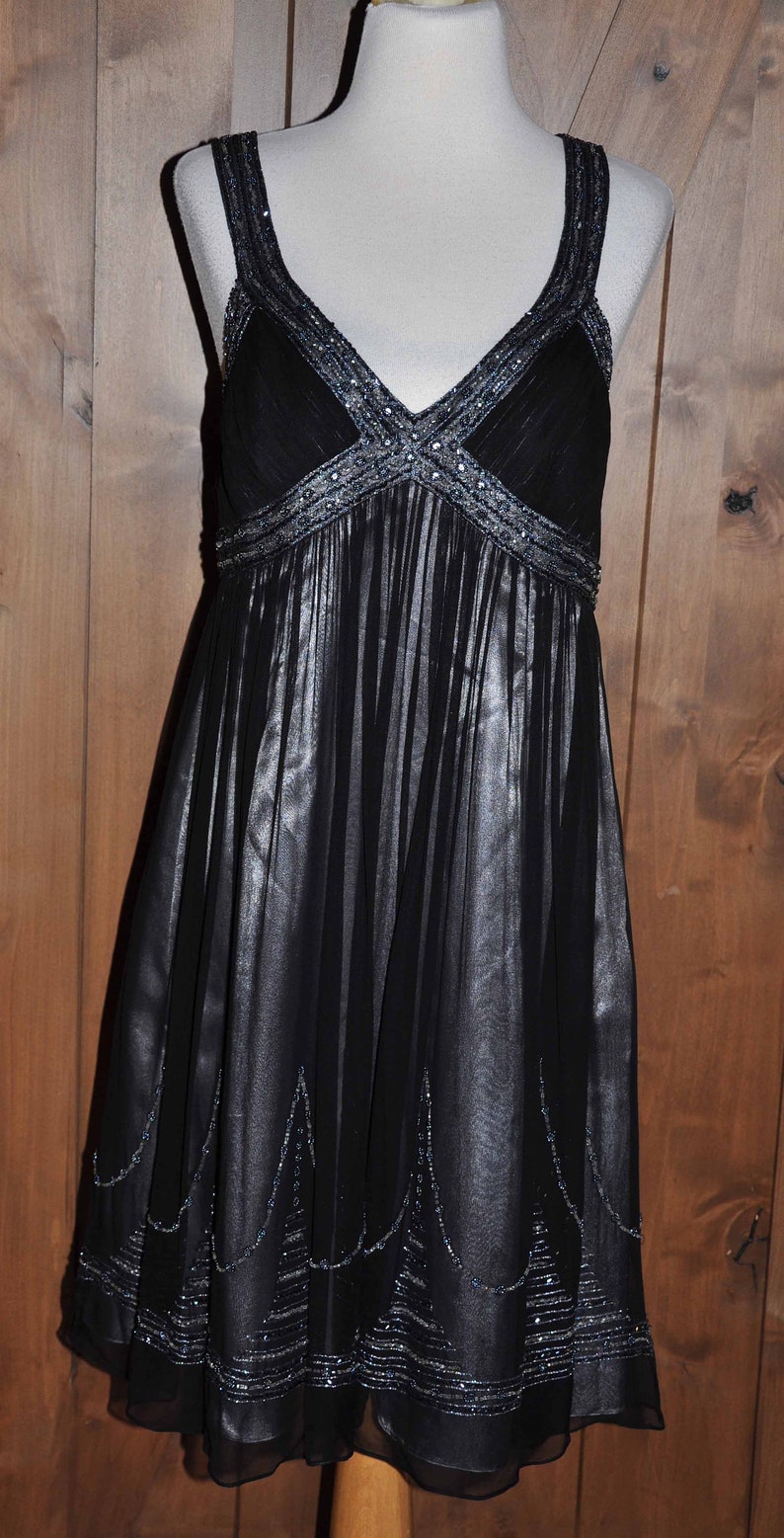 Puede incluir: Vestido de noche negro sobre un maniqu&iacute;. El vestido presenta un escote en V y tirantes adornados con cuentas plateadas. La falda es plisada y tiene un dobladillo con cuentas. El vestido es de tejido transparente.