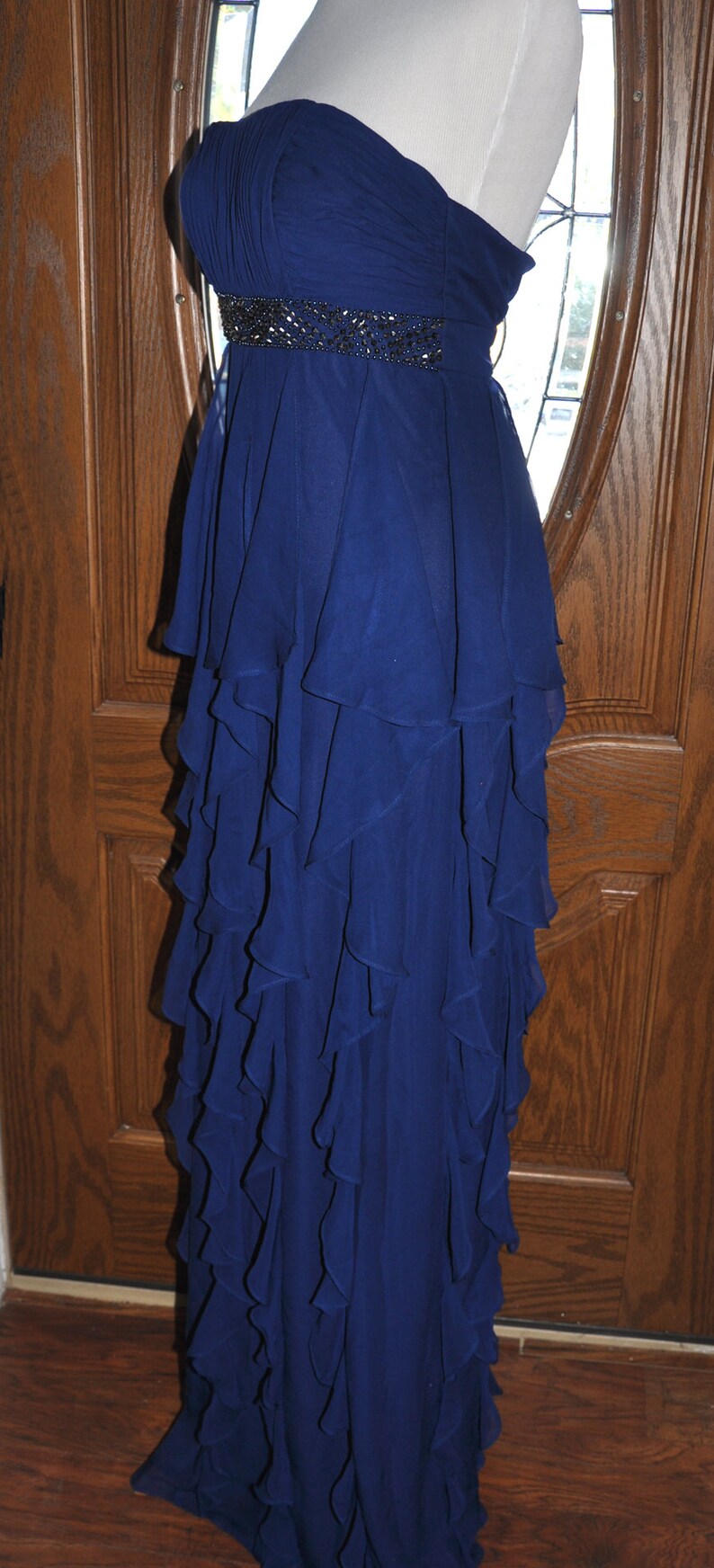 Puede incluir: Un vestido de noche azul marino sin tirantes con un cintur&oacute;n de cuentas. El vestido presenta una falda con volantes en capas. El corpi&ntilde;o est&aacute; fruncido y el vestido se exhibe en un maniqu&iacute;. El vestido mide aproximadamente 152 cm de largo.