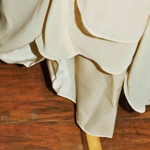 K&ouml;nnte beinhalten: Nahaufnahme des Saums eines wei&szlig;en, flie&szlig;enden Kleides mit einem R&uuml;schen-Saum. Das Kleid h&auml;ngt an einem Holzb&uuml;gel.