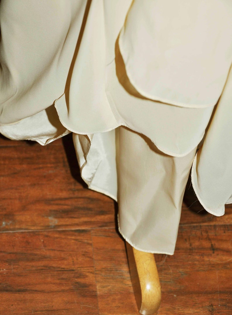 K&ouml;nnte beinhalten: Nahaufnahme des Saums eines wei&szlig;en, flie&szlig;enden Kleides mit einem R&uuml;schen-Saum. Das Kleid h&auml;ngt an einem Holzb&uuml;gel.