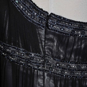 Puede incluir: Primer plano de un vestido negro con detalles de cuentas en el cuello y la cintura. El vestido tiene una tela transparente y fluida con l&iacute;neas verticales, y las cuentas son en tonos plateados y azul oscuro.