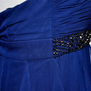 Puede incluir: Un vestido de fiesta azul oscuro con un solo hombro. El corpi&ntilde;o presenta tela fruncida y un cintur&oacute;n con cuentas. La falda fluye hacia afuera desde la cintura. El vestido se exhibe en un maniqu&iacute;.