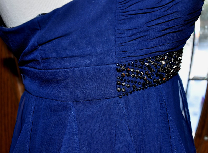 Puede incluir: Un vestido de fiesta azul oscuro con un solo hombro. El corpi&ntilde;o presenta tela fruncida y un cintur&oacute;n con cuentas. La falda fluye hacia afuera desde la cintura. El vestido se exhibe en un maniqu&iacute;.