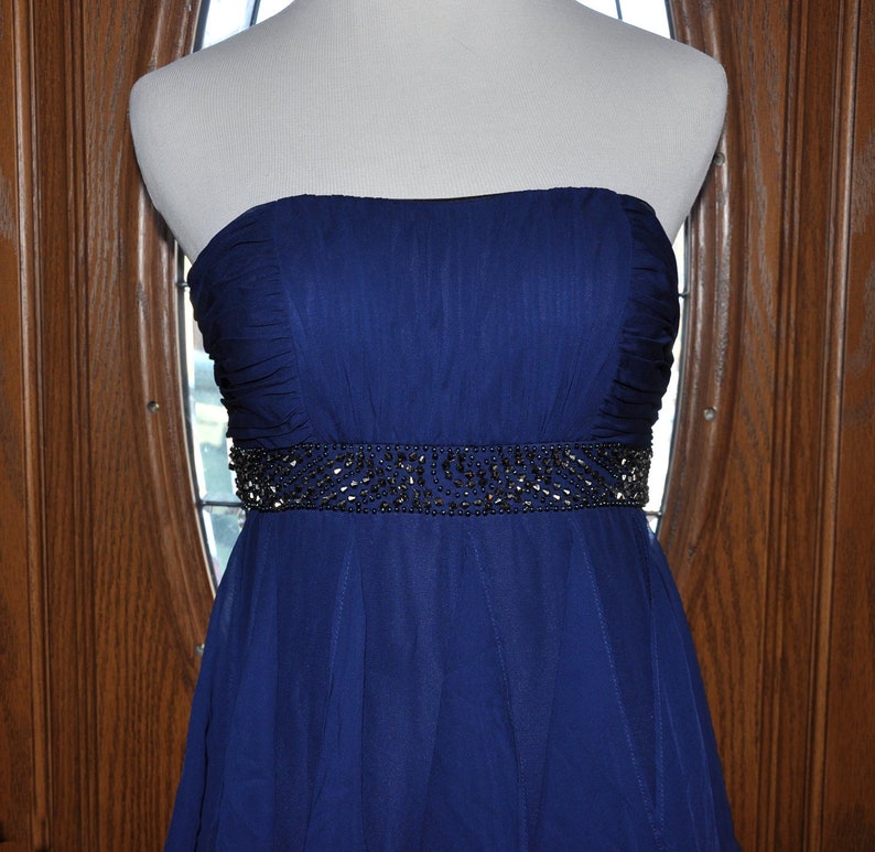 Puede incluir: Un vestido de fiesta azul marino sin tirantes. El vestido presenta un cintur&oacute;n con cuentas y una falda fluida. El corpi&ntilde;o est&aacute; fruncido y el vestido se exhibe en un maniqu&iacute;. El fondo es una puerta de madera con una ventana.