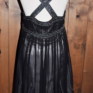 Puede incluir: Vestido de noche negro con falda fluida y tirantes cruzados. Los tirantes y la cintura est&aacute;n adornados con abalorios plateados. El vestido se exhibe en un maniqu&iacute; sobre un fondo de madera.