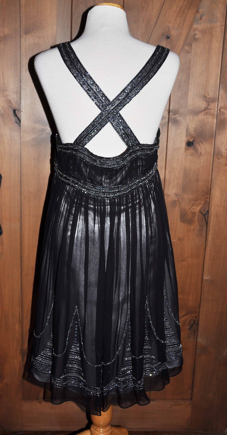 Puede incluir: Vestido de noche negro con falda fluida y tirantes cruzados. Los tirantes y la cintura est&aacute;n adornados con abalorios plateados. El vestido se exhibe en un maniqu&iacute; sobre un fondo de madera.