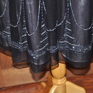 Puede incluir: Vestido negro con adornos de cuentas y lentejuelas. El tejido transparente del vestido presenta l&iacute;neas de cuentas plateadas y azules a lo largo del dobladillo y en cascada desde el escote. El vestido se exhibe en un maniqu&iacute; de madera.
