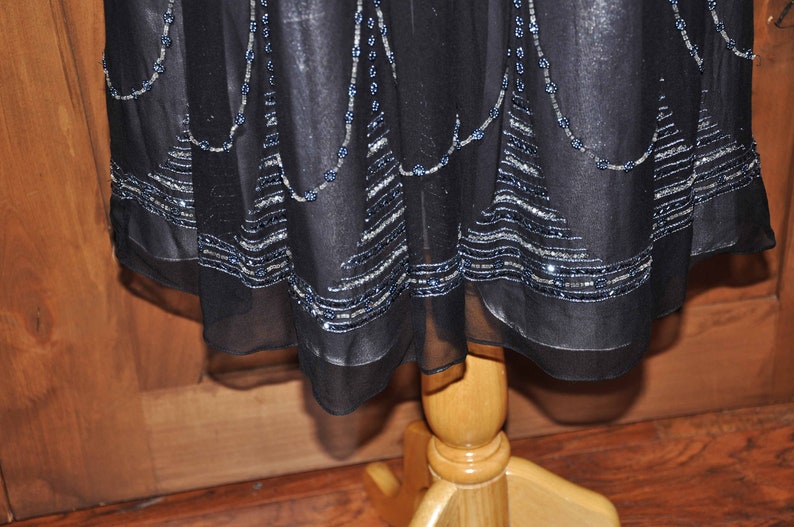 Puede incluir: Vestido negro con adornos de cuentas y lentejuelas. El tejido transparente del vestido presenta l&iacute;neas de cuentas plateadas y azules a lo largo del dobladillo y en cascada desde el escote. El vestido se exhibe en un maniqu&iacute; de madera.