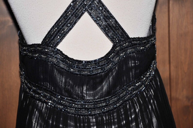 Puede incluir: Vestido de noche negro con cuello halter y tirantes cruzados en la espalda. Los tirantes y el corpi&ntilde;o est&aacute;n adornados con abalorios plateados. El vestido tiene una falda fluida y plisada, creando una silueta elegante.