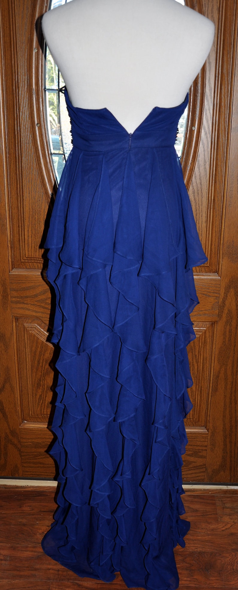Puede incluir: Vestido de noche azul real sin tirantes, largo hasta el suelo, con falda con volantes en capas. El vestido se exhibe en un maniqu&iacute;, mostrando su elegante dise&ntilde;o y tejido fluido. La parte trasera del vestido tiene cierre de cremallera.