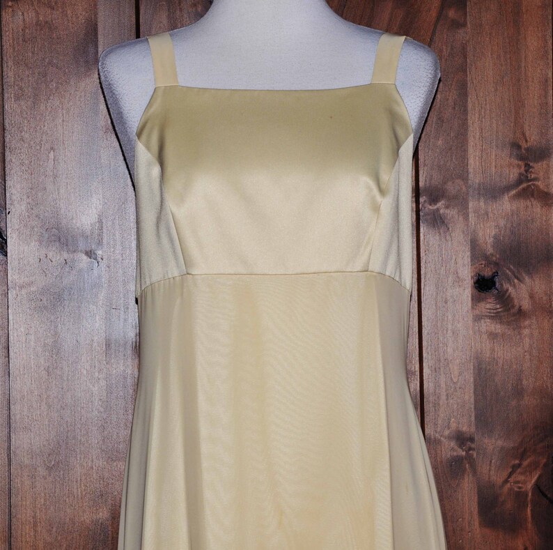 K&ouml;nnte beinhalten: Ein schlichtes, &auml;rmelloses, bodenlanges Kleid aus hellem gelbem Satin. Das Kleid hat einen quadratischen Ausschnitt und ein tailliertes Oberteil.