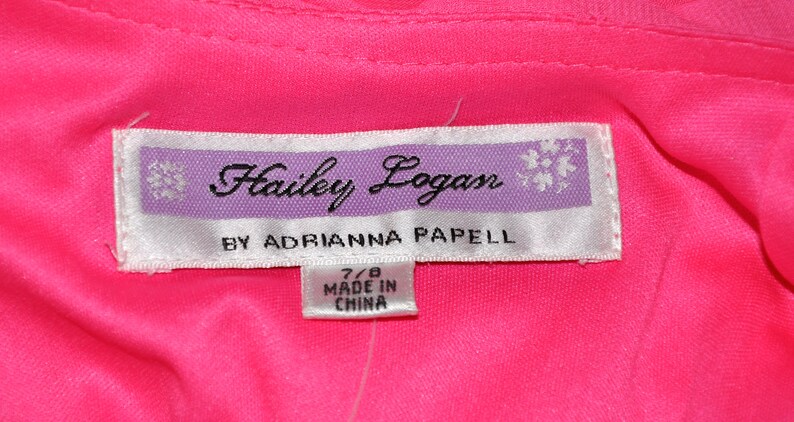 以下が含まれることがあります： 紫色の縁取りのある白いタグに「Hailey Logan by Adrianna Papell」という文字と、「7/8 Made in China」と書かれた小さなタグが付いている。