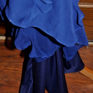 Puede incluir: Un vestido azul cobalto en capas con una parte superior fluida con volantes y una falda s&oacute;lida m&aacute;s oscura. El vestido est&aacute; colgado de una percha de madera, mostrando la textura y la ca&iacute;da de la tela. El vestido est&aacute; hecho de un material suave y fluido.