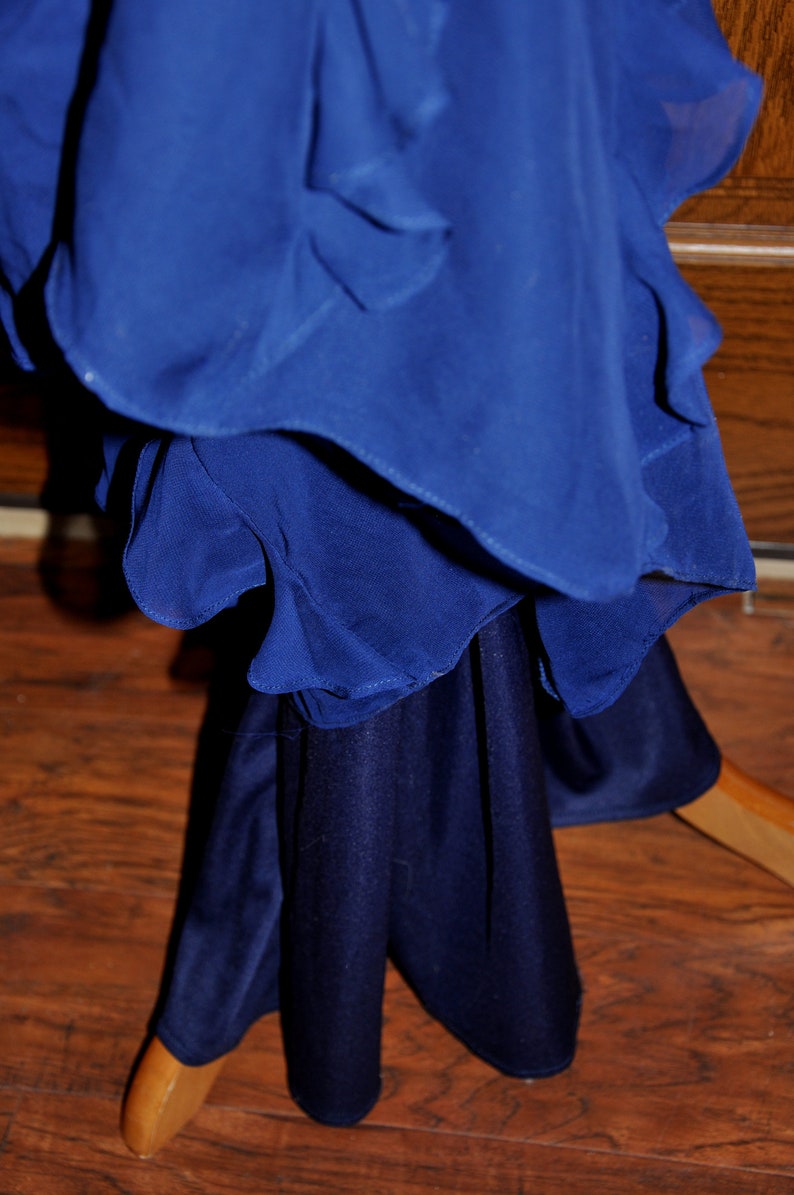 Puede incluir: Un vestido azul cobalto en capas con una parte superior fluida con volantes y una falda s&oacute;lida m&aacute;s oscura. El vestido est&aacute; colgado de una percha de madera, mostrando la textura y la ca&iacute;da de la tela. El vestido est&aacute; hecho de un material suave y fluido.