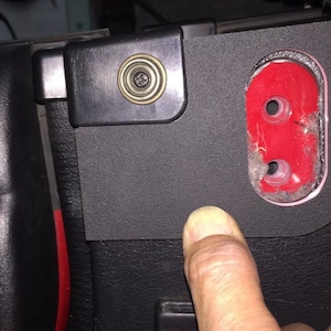 May include: Close-up of a black plastic panel with a red oval cutout. The cutout has two holes in it. The panel is attached to a black leather surface.