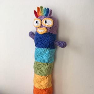 以下が含まれることがあります： 手作りのカラフルなかぎ針編みの人形。奇抜な生き物の形をしており、虹色の鶏冠、黒い瞳の大きな白い目、青、緑、黄、オレンジ、赤の色合いのセグメント化された体を持っています。小さな腕と足がデザインを完成させています。